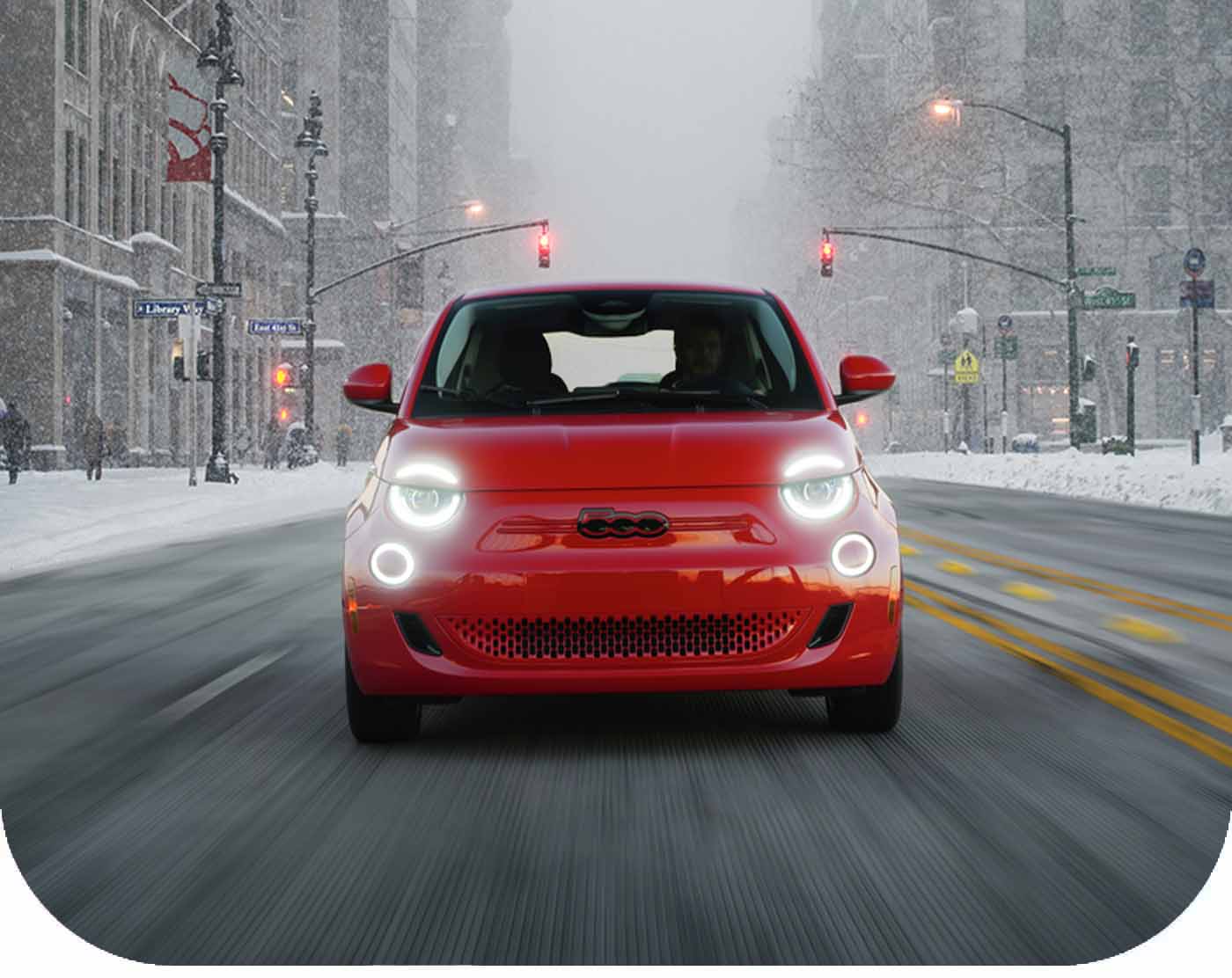 A head-on angle of a 2024 Fiat 500e traveling down a city street in winter during a snowstorm.