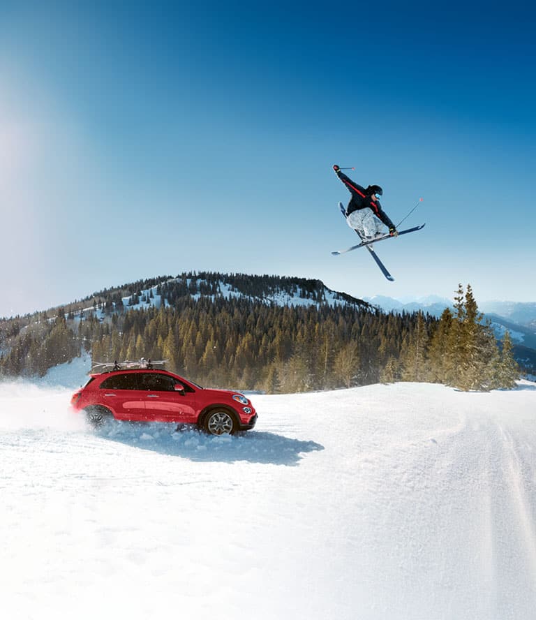 A passenger-side profile of a red 2022 Fiat 500X Trekking with aftermarket equipment, being driven through heavy snow, kicking up the white powder as it goes, while a skier jumps overhead and mountains rise in the background.