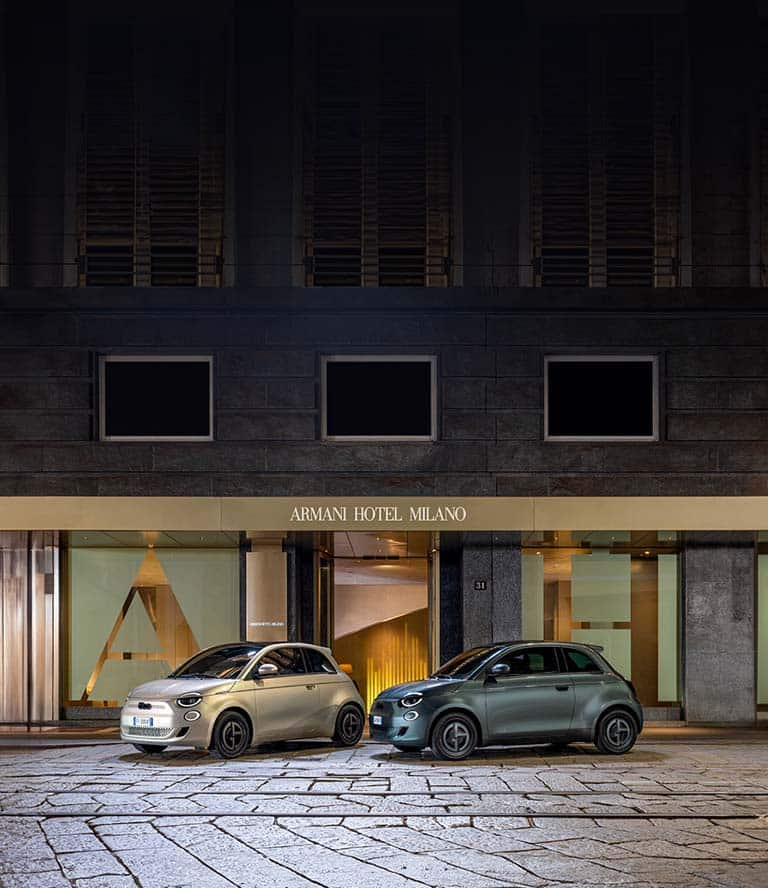 Two 2025 Fiat 500e Giorgio Armani Collector's Edition models parked on a cobblestone street. On the left a model in gray beige and on the right, a model in dark green.