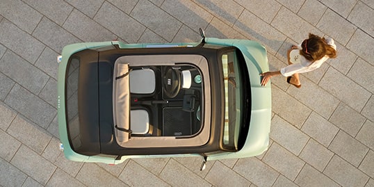 A bird's-eye view of a mint green Fiat Topolino with its sunroof open and a woman patting the hood of the vehicle.
